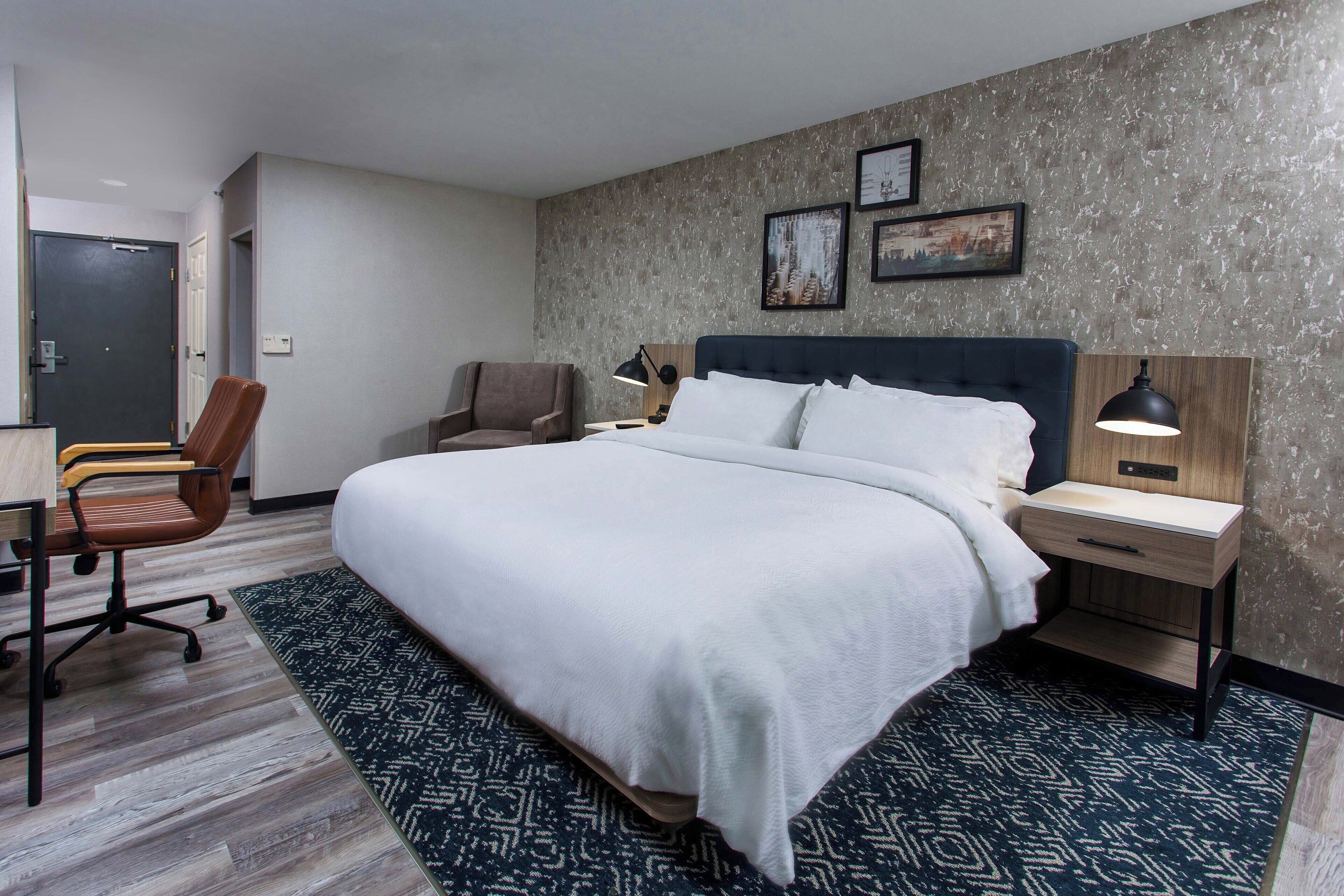 A hotel room with a large bed, a desk, a chair, and a nightstand with a lamp.