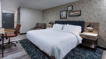 A hotel room with a large bed, a desk, a chair, and a nightstand with a lamp.