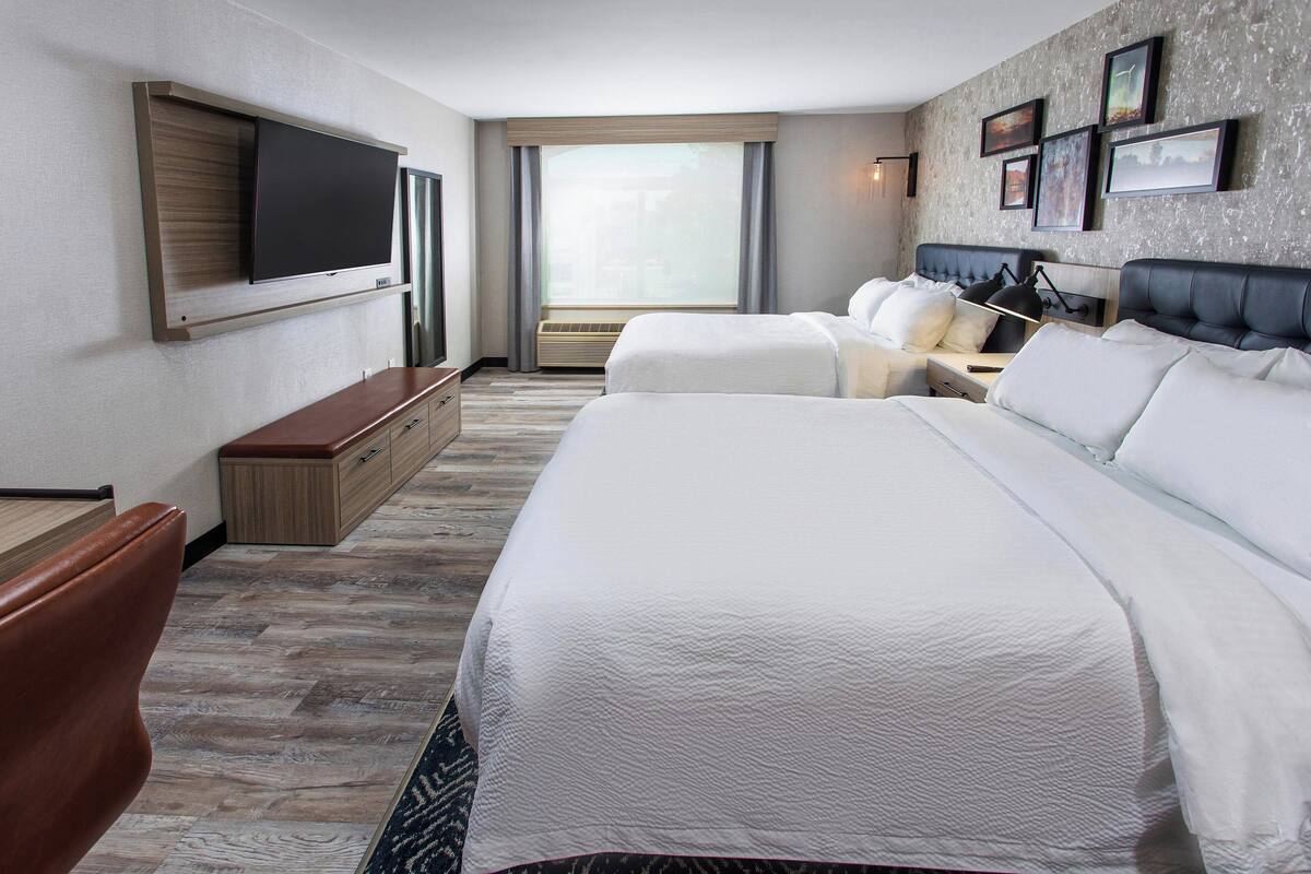 A hotel room with two beds, a flat-screen TV mounted on the wall, a desk, and a window with curtains.