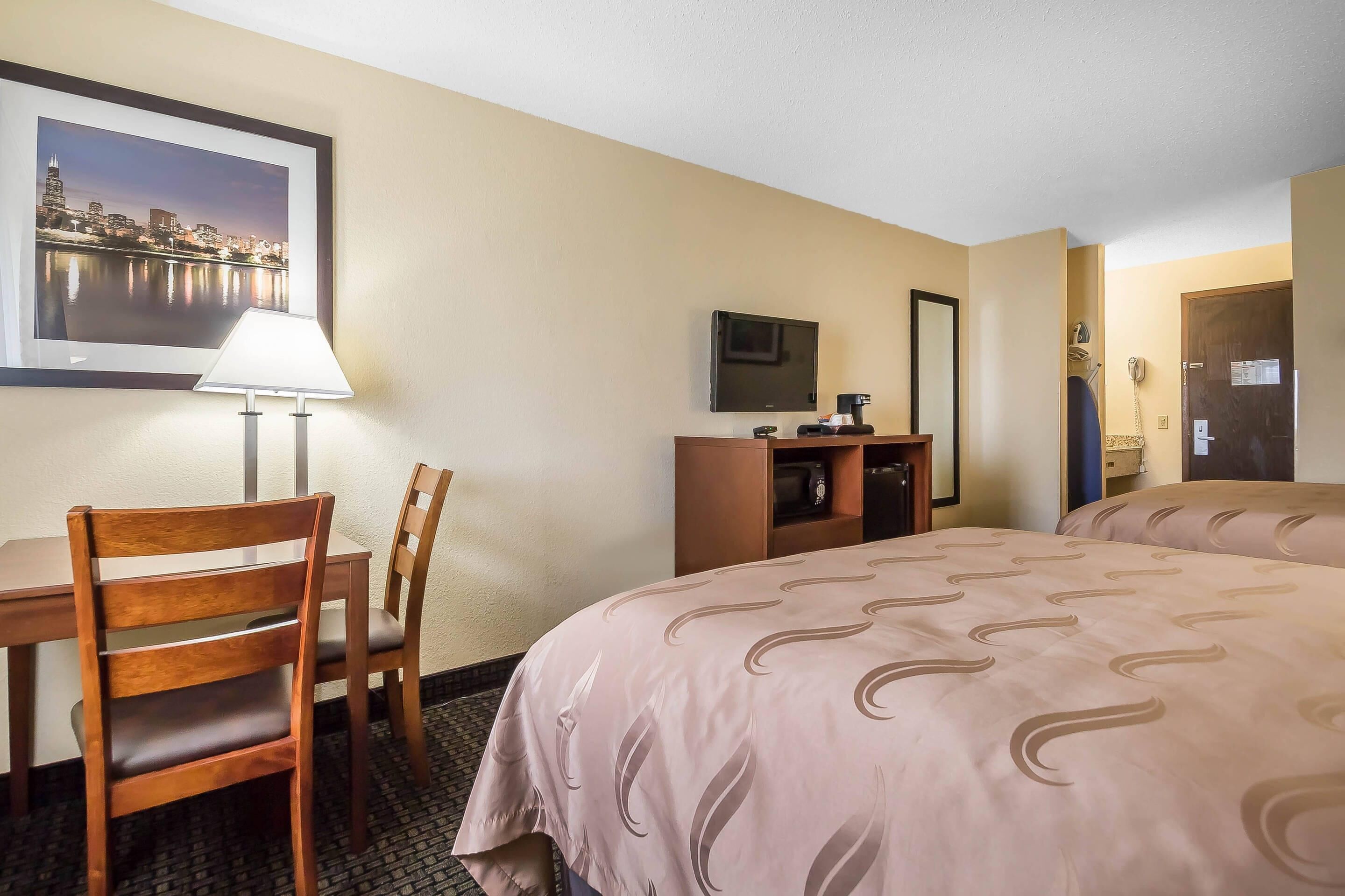 A hotel room with a bed, a desk with a chair, a TV, a lamp, and a framed picture on the wall.