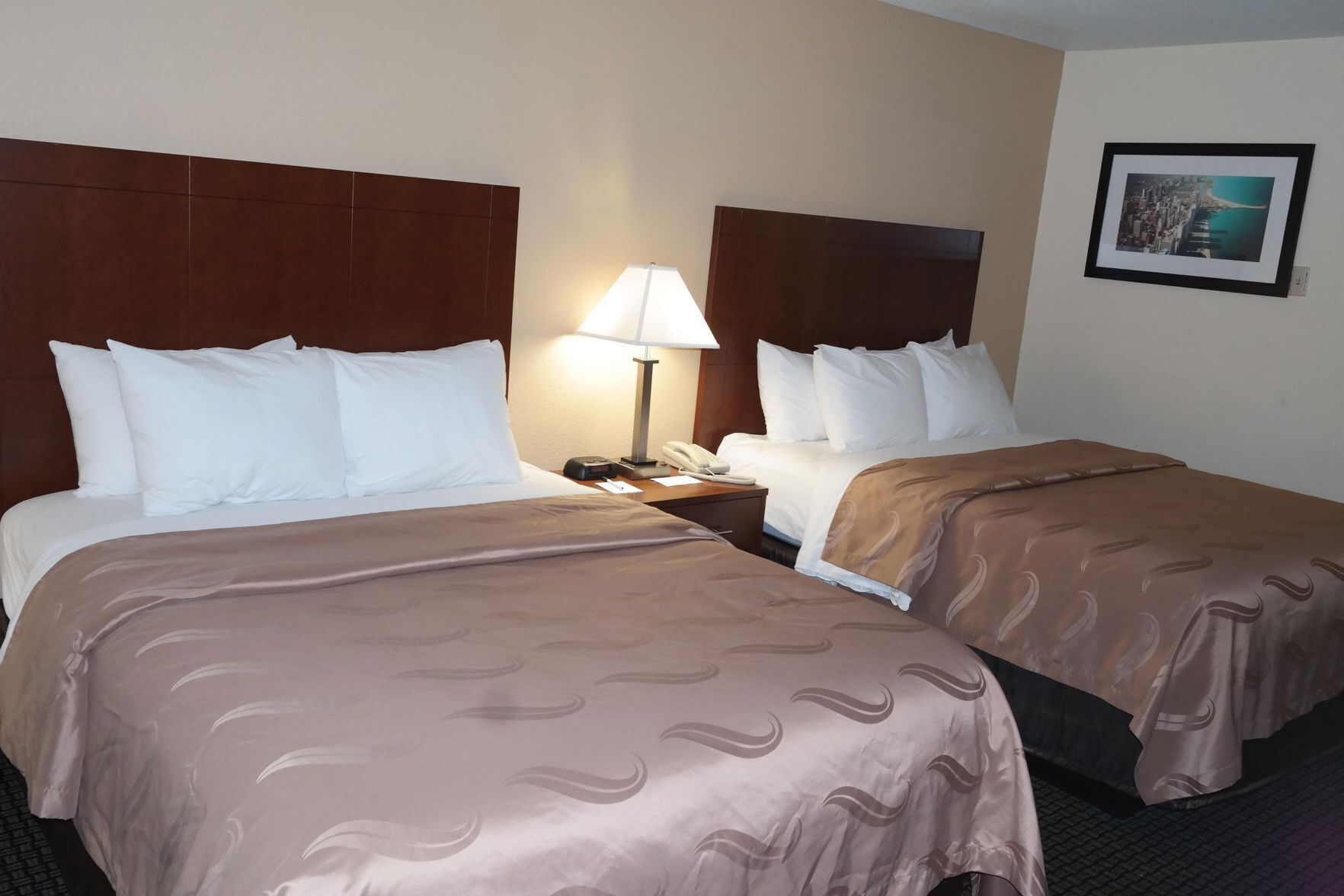 A hotel room with two beds, a nightstand with a lamp, a phone, and a framed picture on the wall.