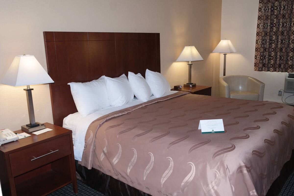 A hotel room with a bed, two bedside lamps, a chair, a desk with a phone, and a wall-mounted air conditioner.