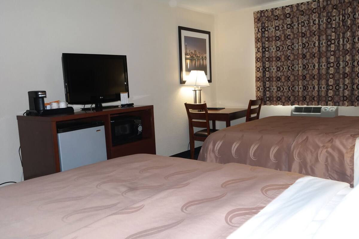 A hotel room with two beds, a TV, a desk with a lamp, a microwave, and a small refrigerator.