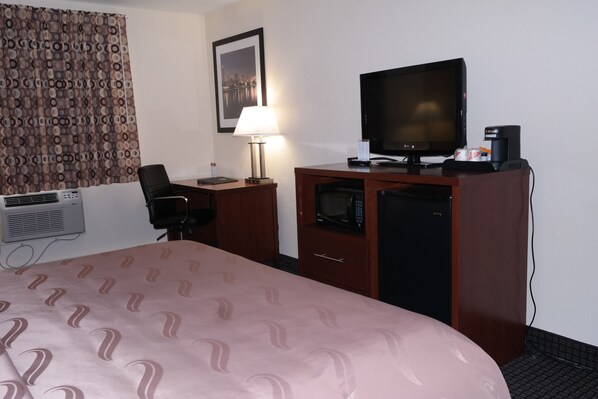 A hotel room with a bed, a desk with a computer, a television, a microwave, and an air conditioning unit.
