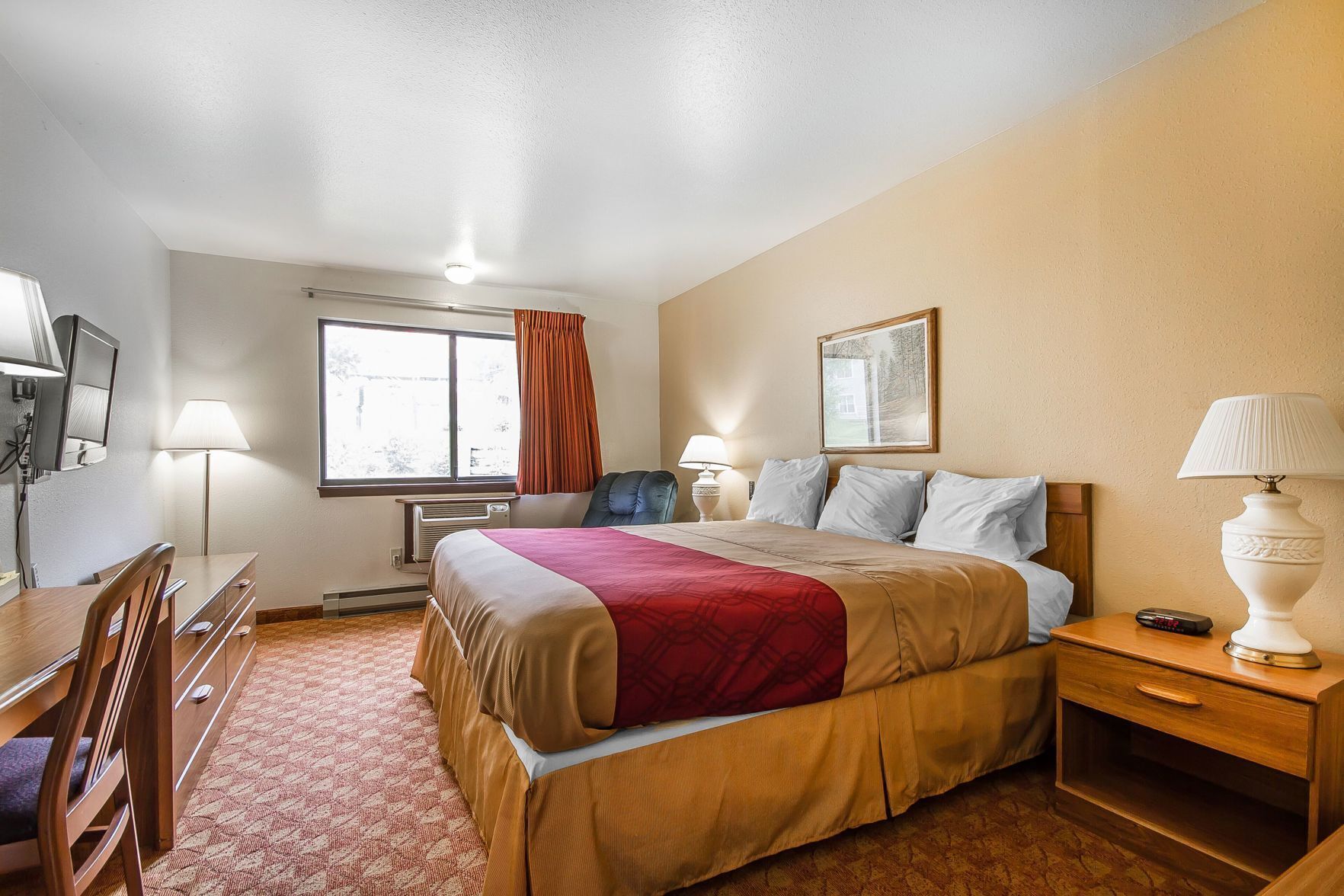 Habitación de hotel con cama, mesitas de noche, escritorio, silla, lámpara, televisor y ventana con cortinas.