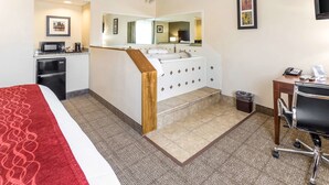 A hotel room with a bed, a desk, a chair, a bathroom with a sink and mirror, a microwave, and a trash can.