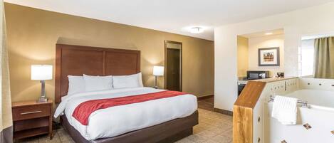 Quarto de hotel com uma cama grande, cabeceira de madeira, escrivaninha com luminária, banheiro com banheira e um quadro pendurado na parede.