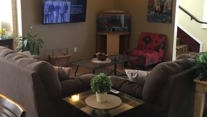 A living room with a flat-screen TV mounted on the wall, a glass coffee table, two sofas, a red armchair, and a small table with a plant. - Cheyenne Guest Inn (Cheyenne)