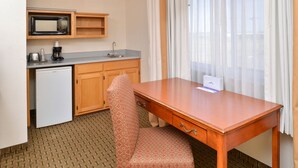 A compact hotel room with a kitchenette, a wooden desk, a patterned chair, and a window with sheer curtains. - Cheyenne Guest Inn (Cheyenne)