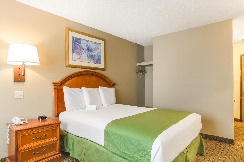 A hotel room with a bed, a bedside table with a lamp, a framed picture on the wall, and a door to the right.