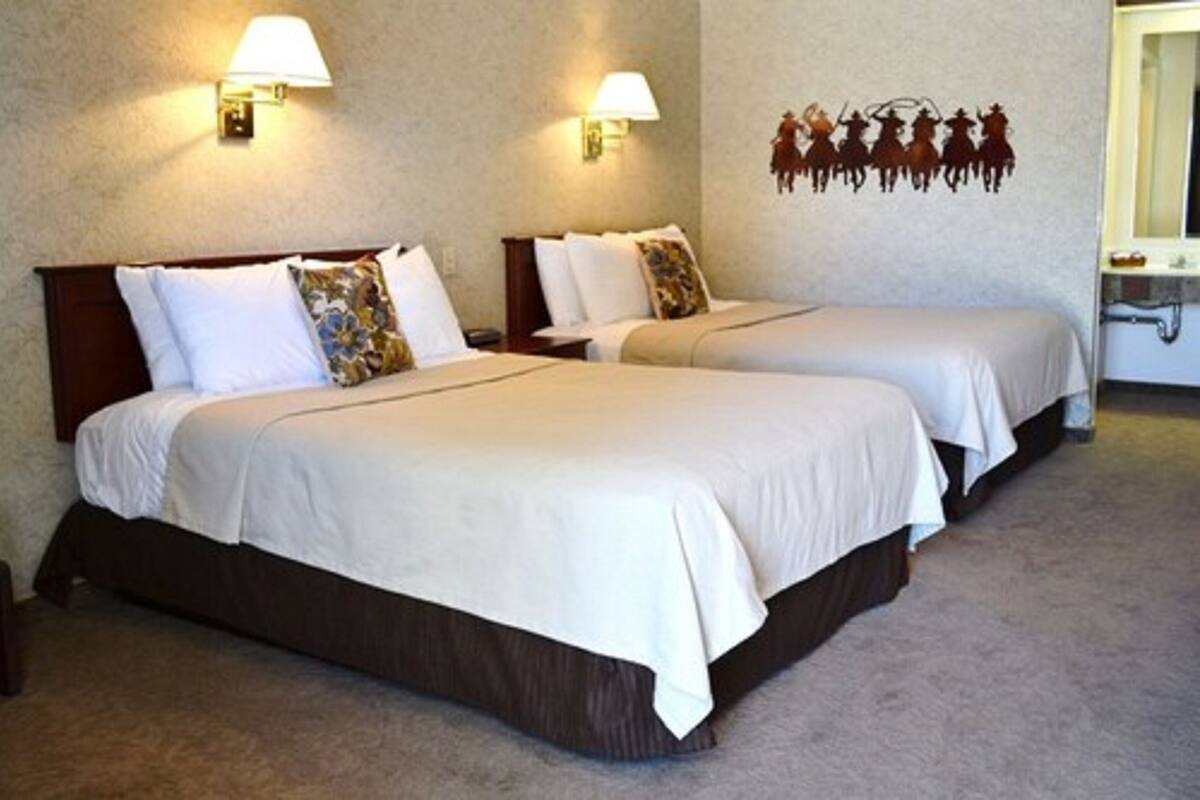 A hotel room with two single beds, each with a wooden headboard, white bedding, and a decorative pillow. There are wall-mounted lamps and a wall decal of cowboys.