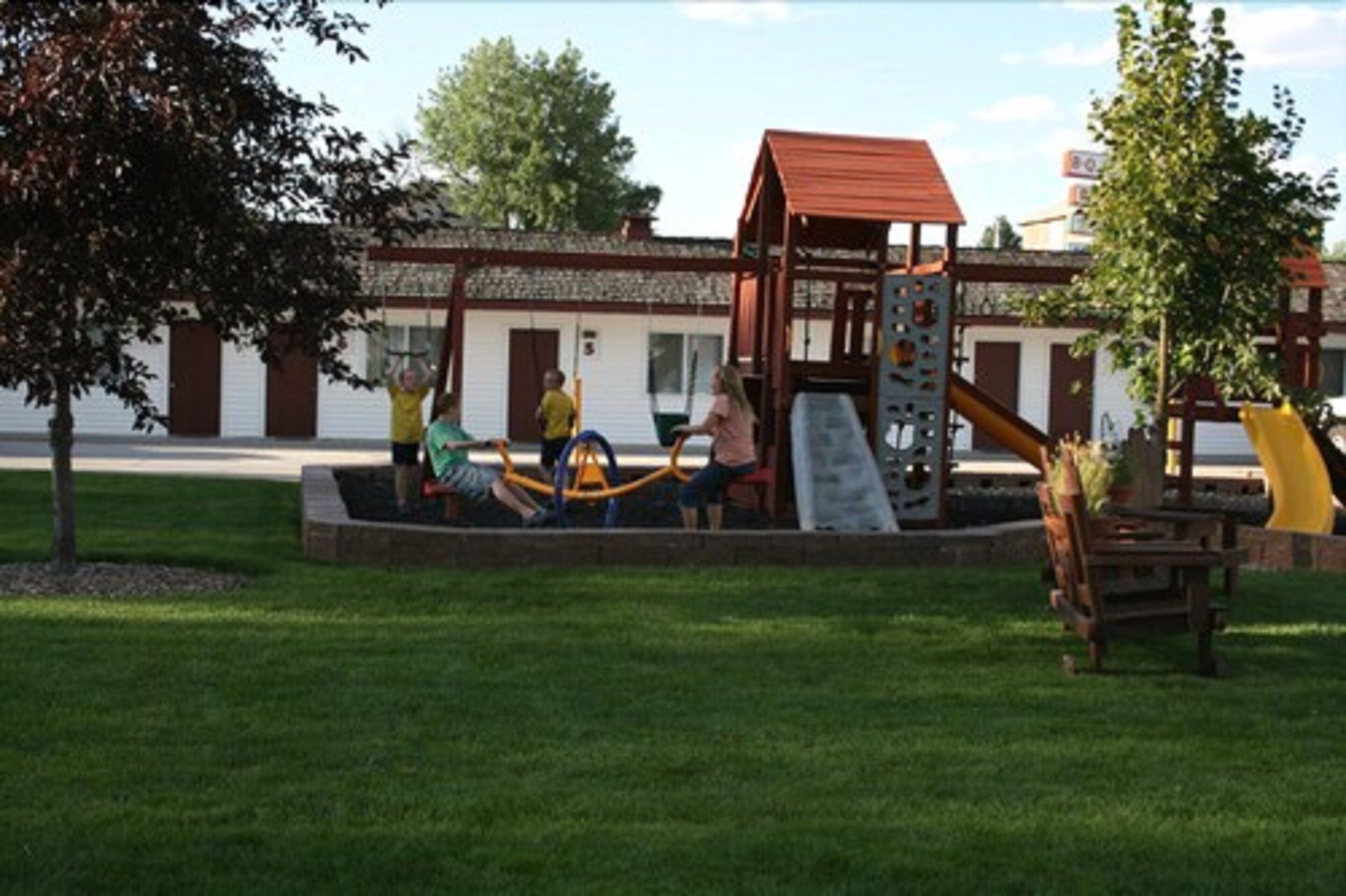 A playground with a slide and climbing structure, surrounded by a grassy area and a building with a red roof.