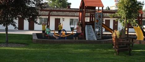 A playground with a slide and climbing structure, surrounded by a grassy area and a building with a red roof.