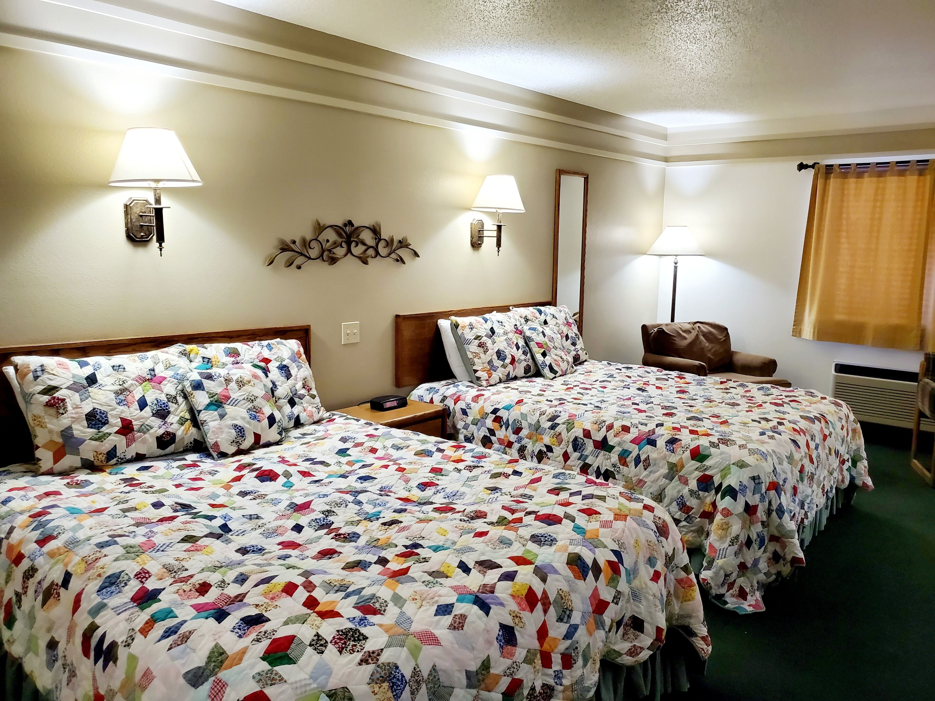 Habitación de hotel con dos camas, cada una con un edredón colorido, lámparas de noche, un espejo y una silla de cuero marrón.