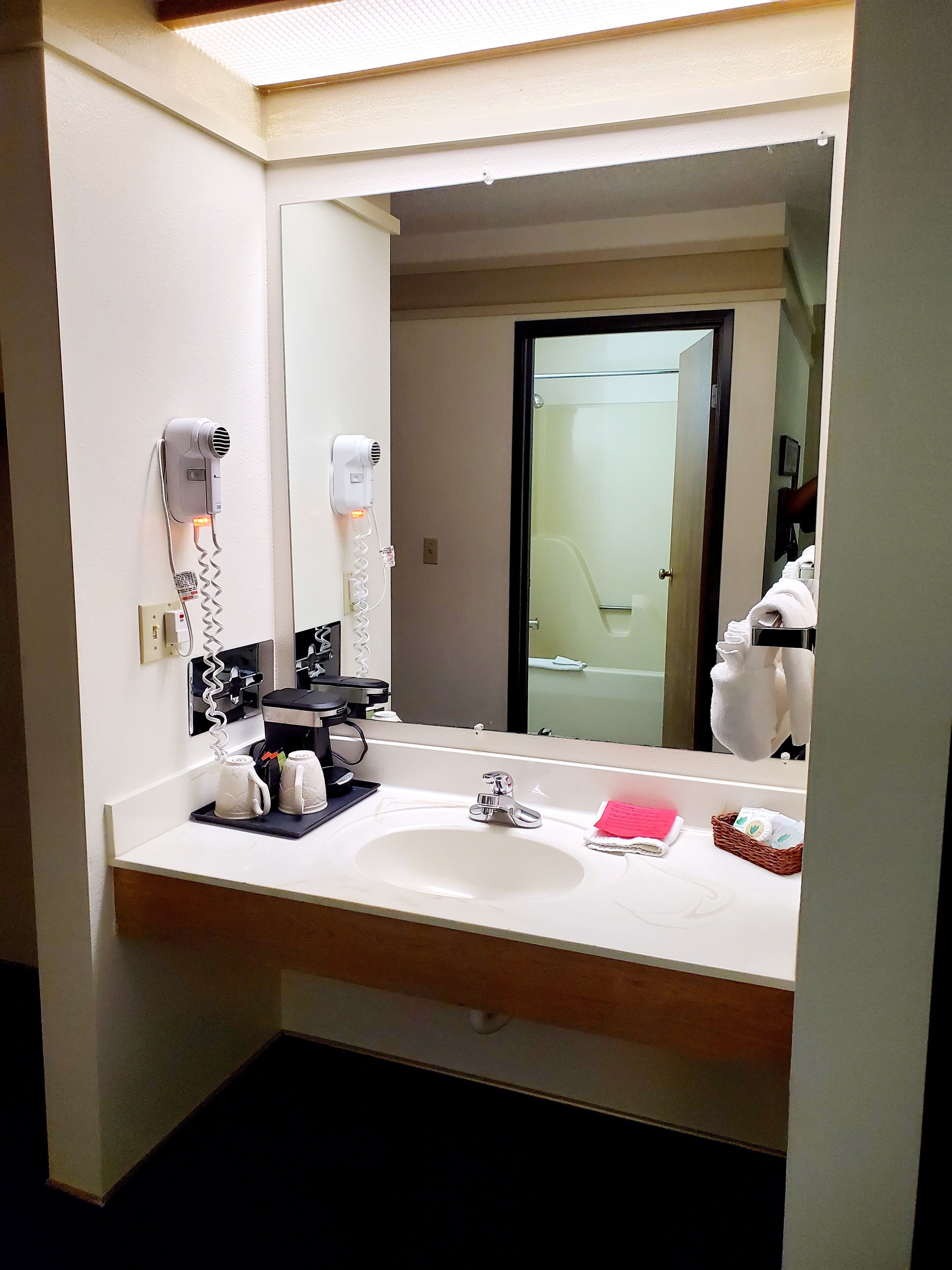 A hotel bathroom with a large mirror, a sink, a hairdryer, and a coffee machine.