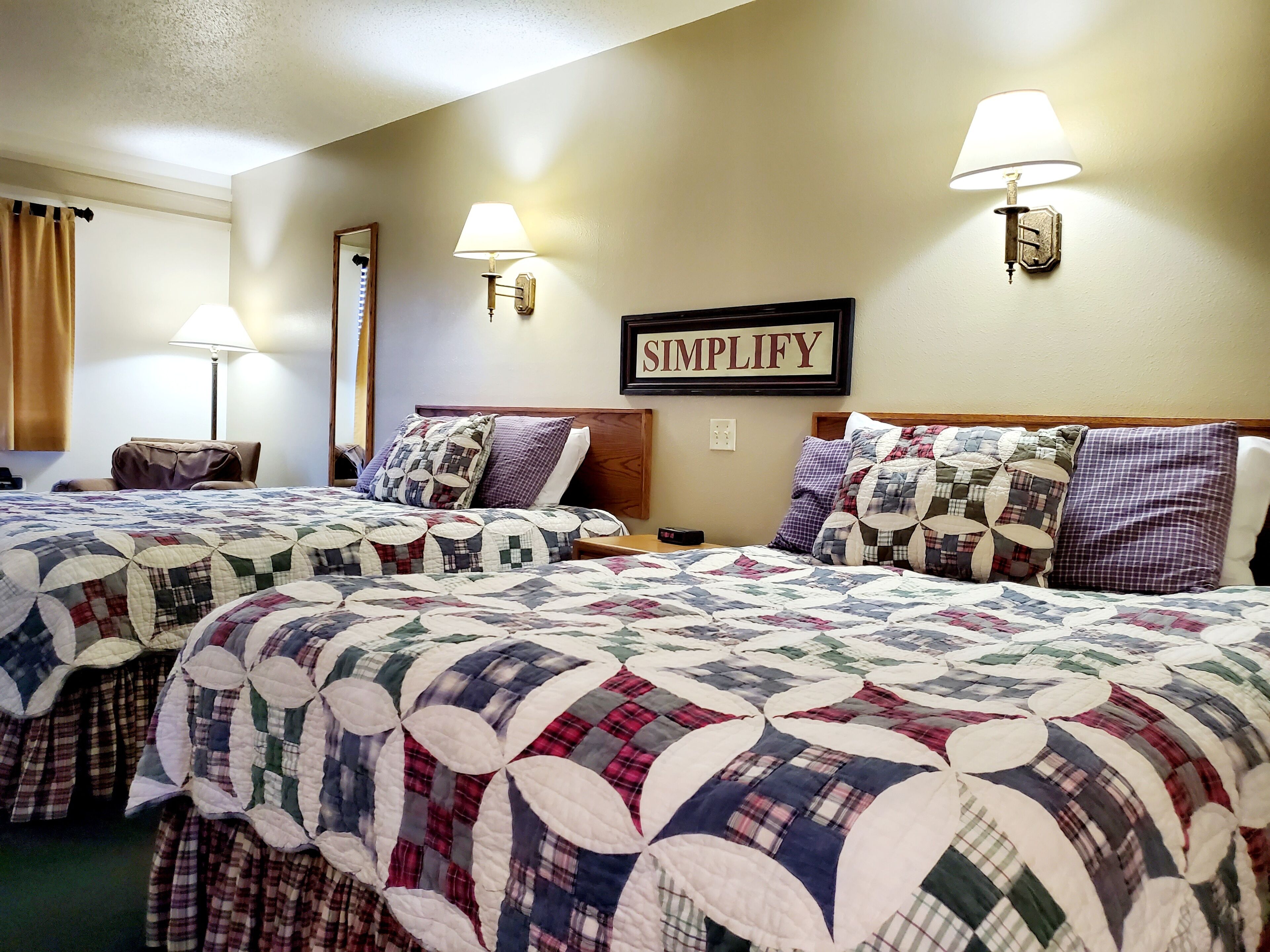 Quarto de hotel com duas camas, cada uma com colcha acolchoada, cabeceira e abajures laterais, e uma placa na parede com a inscrição 'SIMPLIFY'.