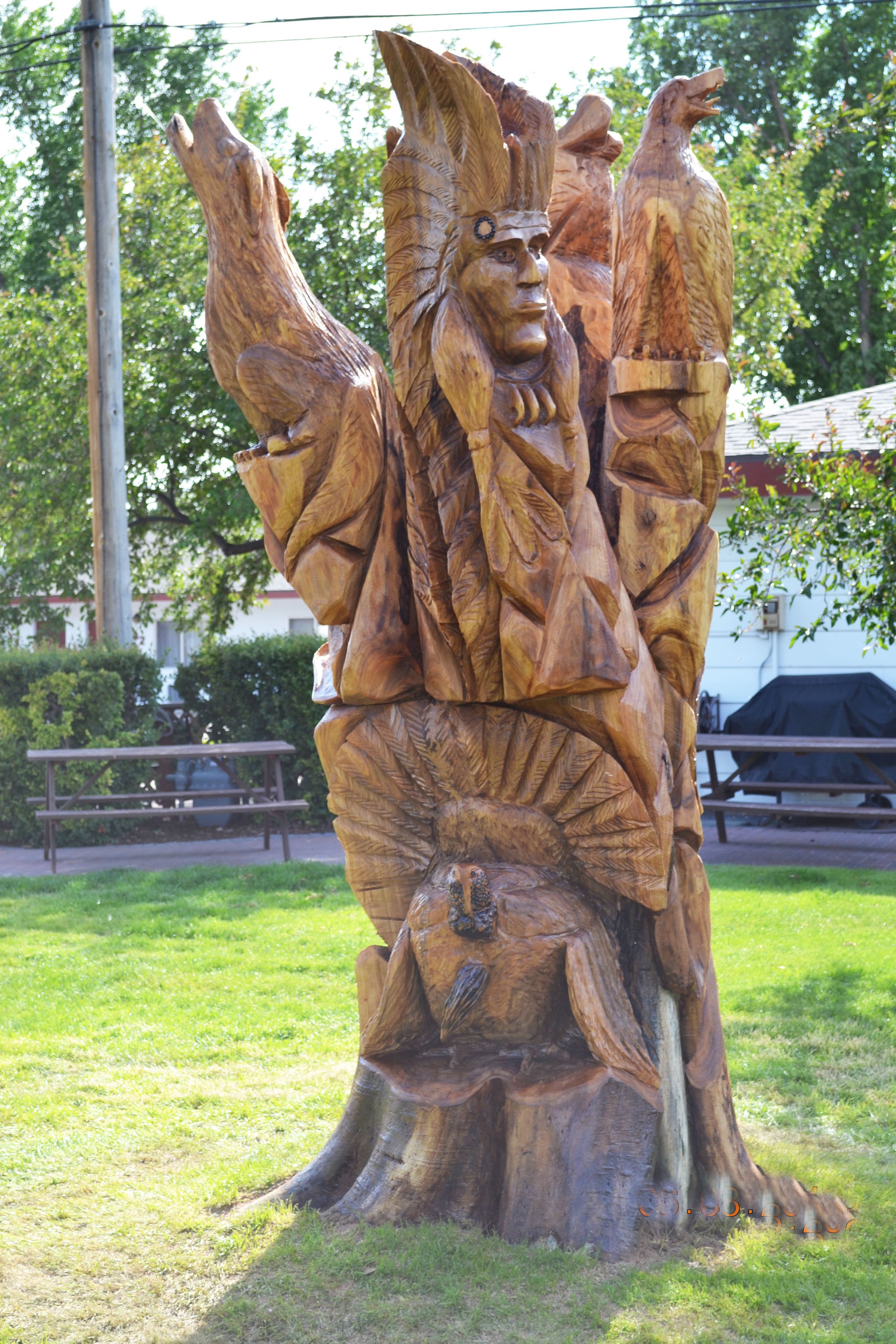 A wooden sculpture of a figure with raised arms, set in a grassy area with a picnic table and benches in the background.