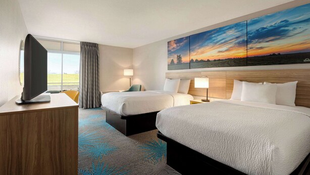 A hotel room with two beds, a TV, a desk, and a large painting of a sunset. - Days Inn by Wyndham Grand Island I-80 (Grand Island)