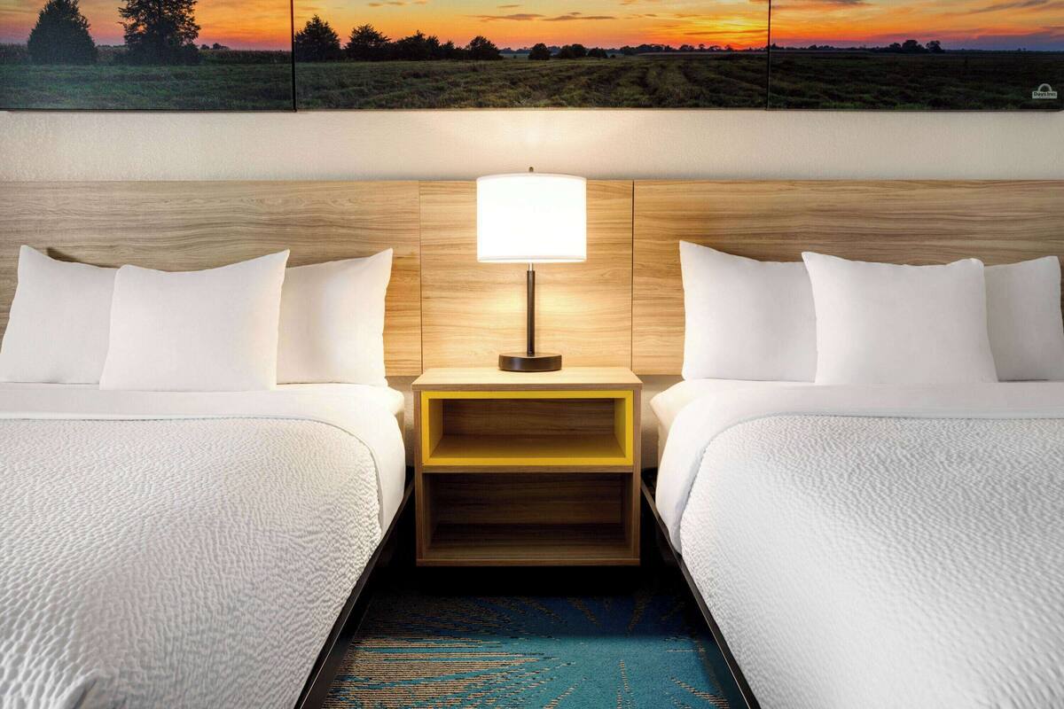 A hotel room with two beds, a nightstand with a lamp, and a large landscape painting above the beds.