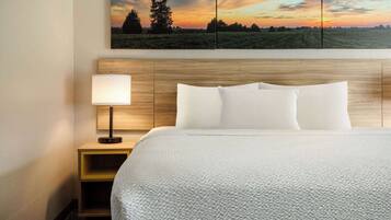 Un cuarto de hotel con una cama grande, una mesita de noche con lámpara y un cuadro colgado en la pared que muestra un paisaje al atardecer.