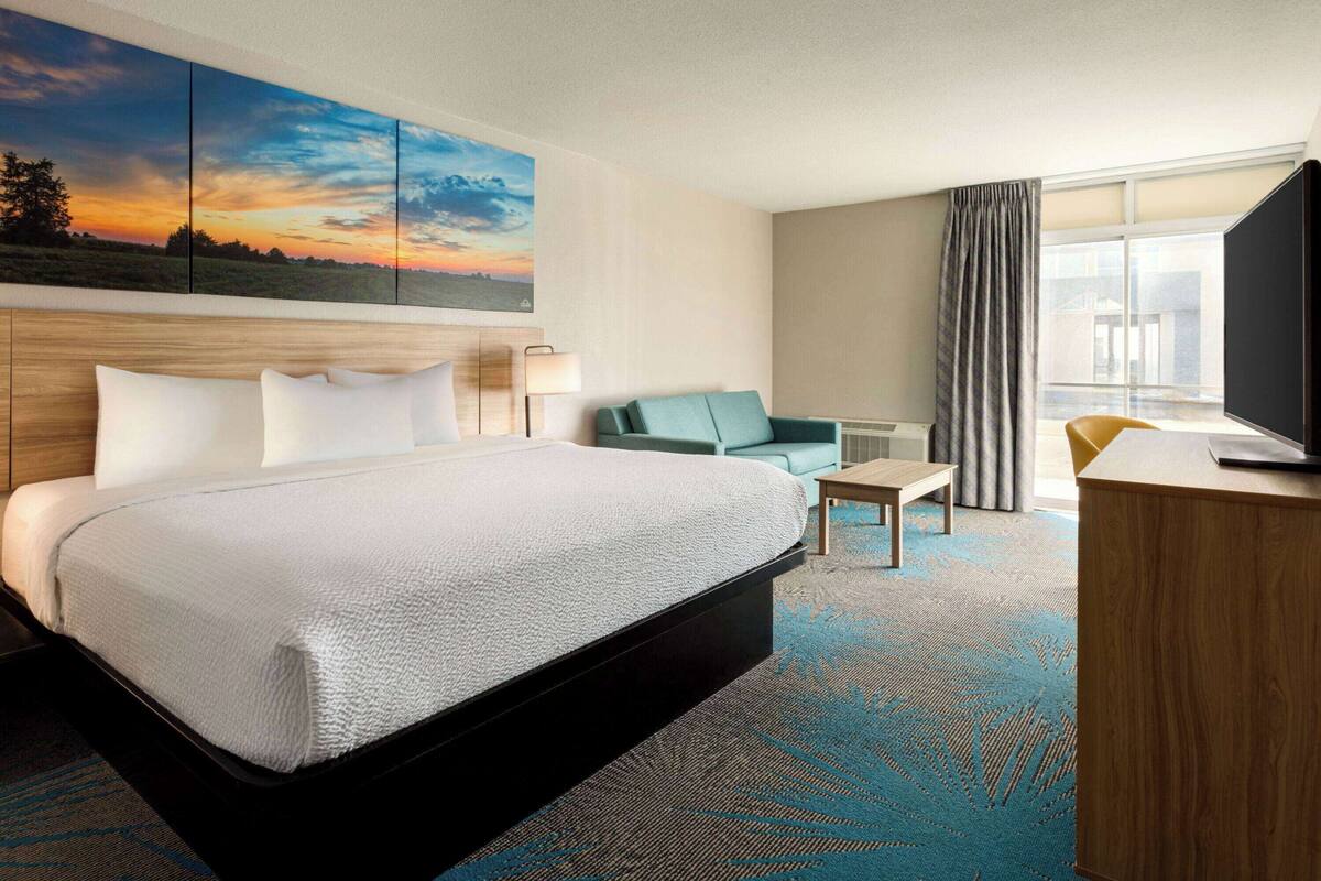 A hotel room with a large bed, a desk, a chair, a TV, and a painting of a sunset on the wall.