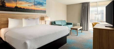A hotel room with a large bed, a desk, a chair, a TV, and a painting of a sunset on the wall.