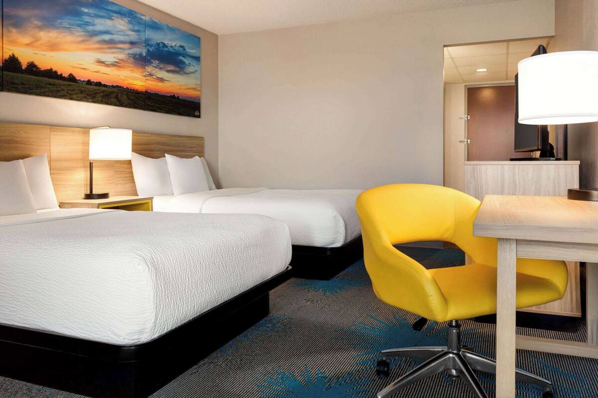 A hotel room with two beds, a desk, a chair, a lamp, and a painting of a sunset.