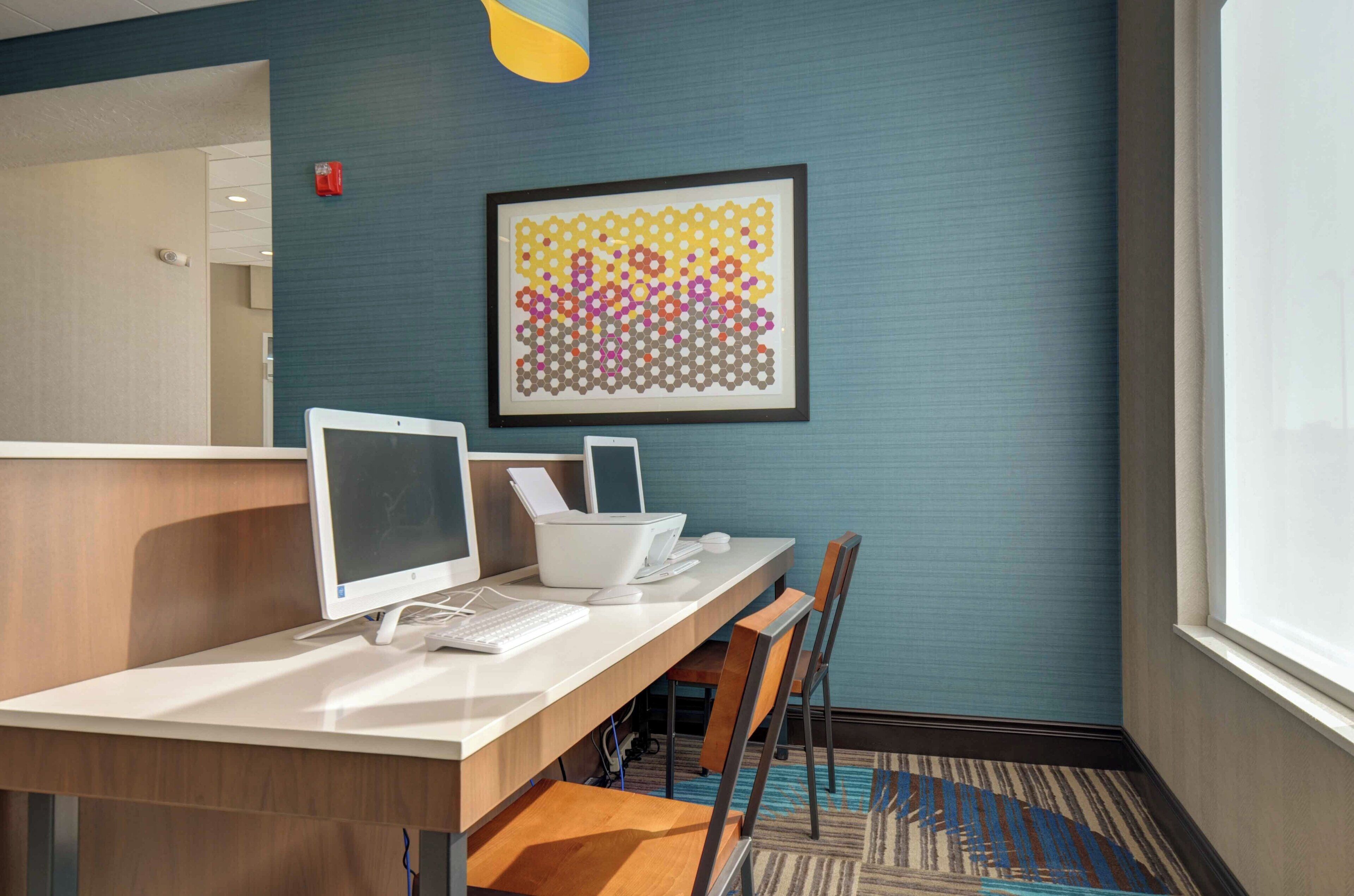 A hotel room with a desk, computer, chair, and a framed abstract art piece on the wall.
