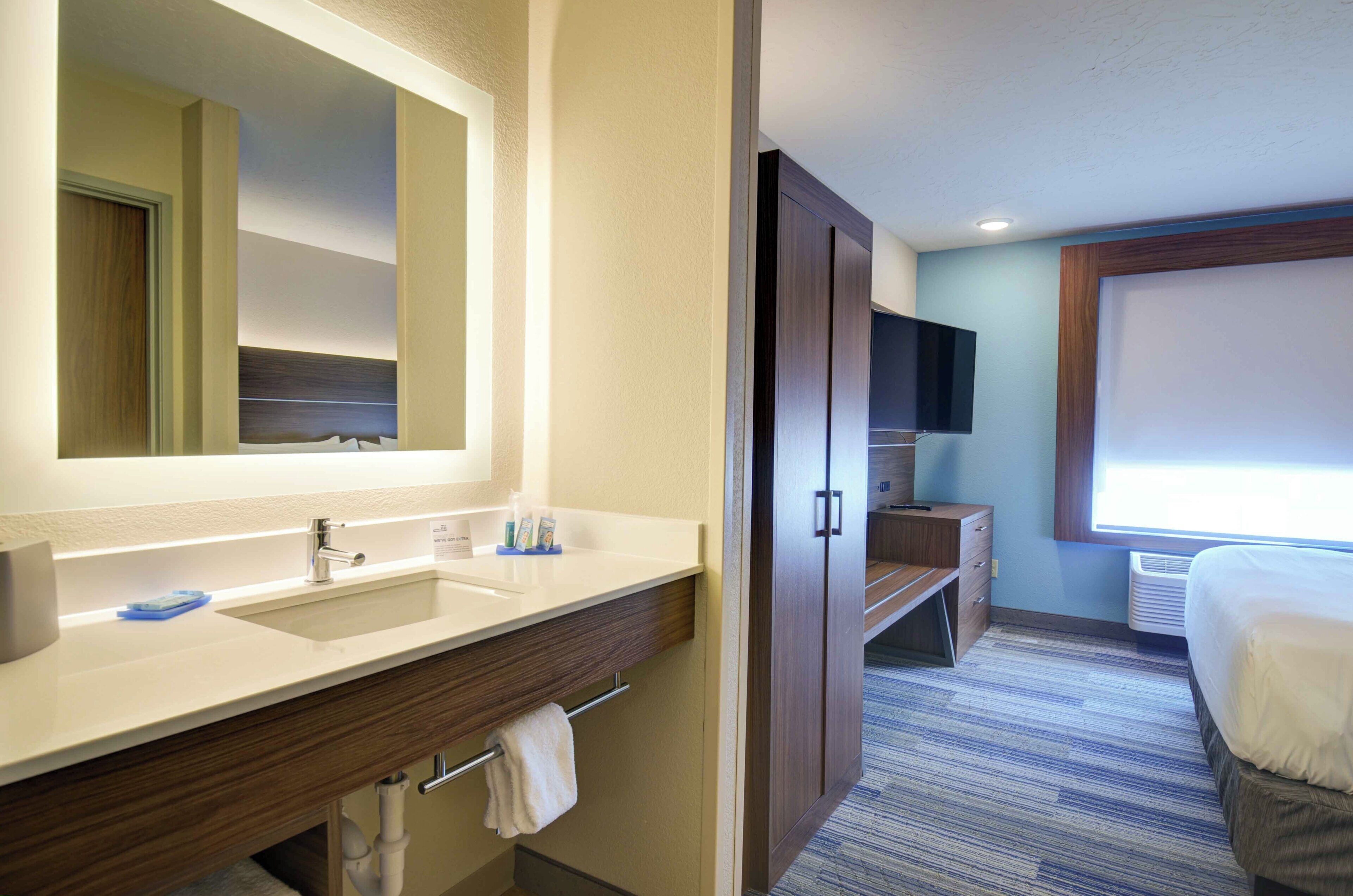 Un baño de hotel con un espejo grande, un lavamanos, un perchero para toallas y un televisor montado en la pared.