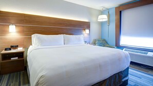 A hotel room with a large bed, a nightstand with a clock, a wall-mounted lamp, and a window with blinds. - Holiday Inn Express & Suites - North Platte by IHG (North Platte)