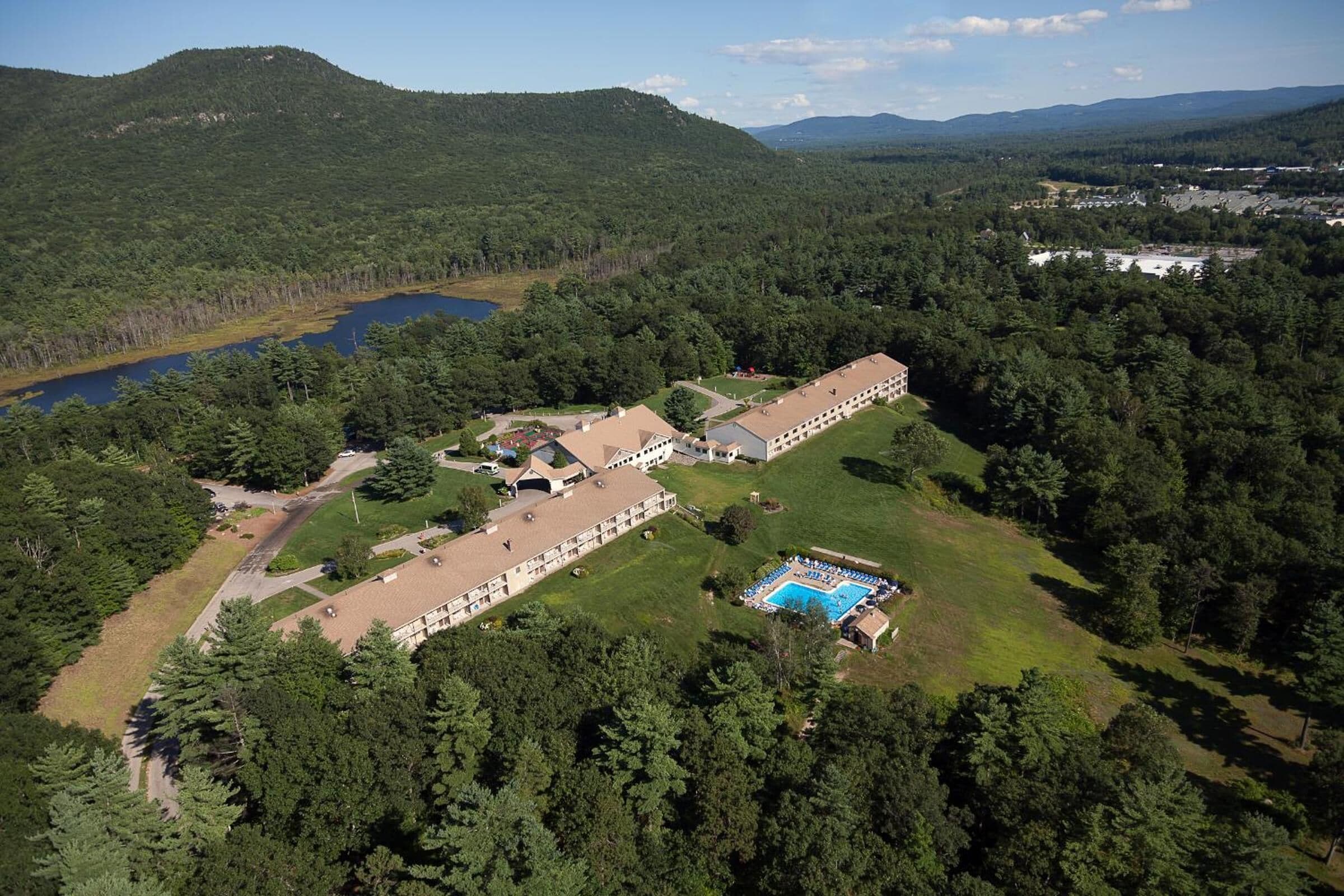 An aerial view of a hotel with a swimming pool, surrounded by dense forest and hills.