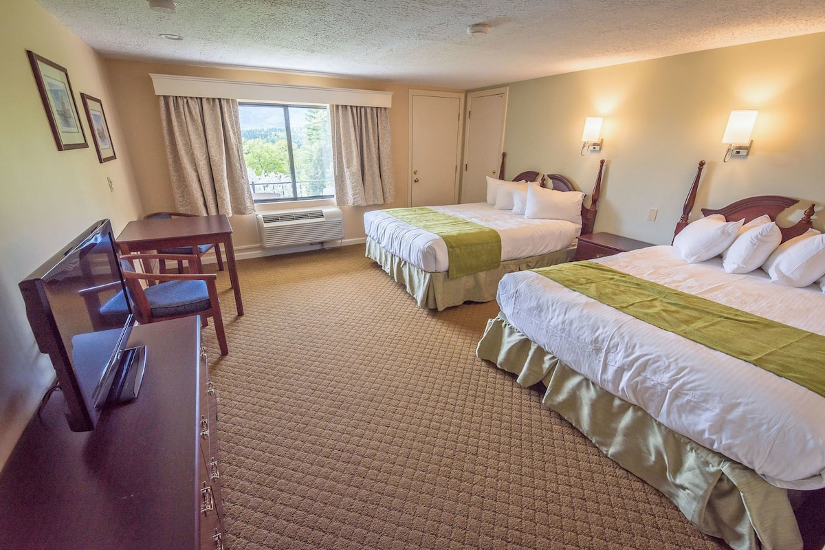 A hotel room with two beds, a desk, and a chair. There is a window with curtains, a wall-mounted heater, and two wall sconces.