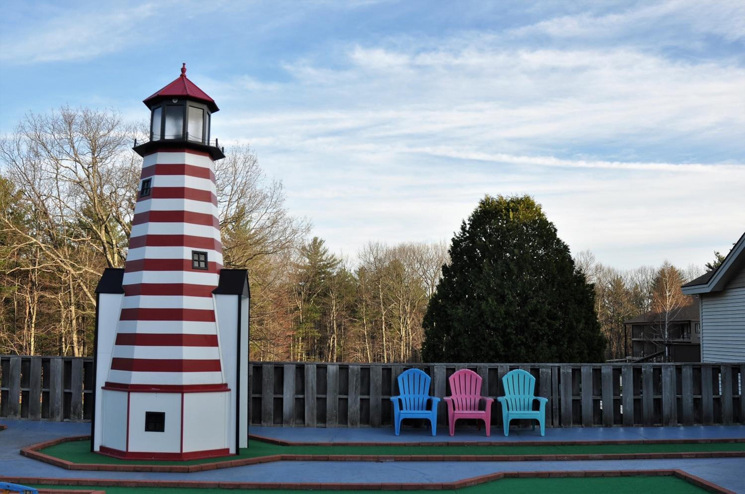 Un faro con una torre rayada de rojo y blanco, una valla de madera y tres sillas Adirondack en azul, rosa y verde.