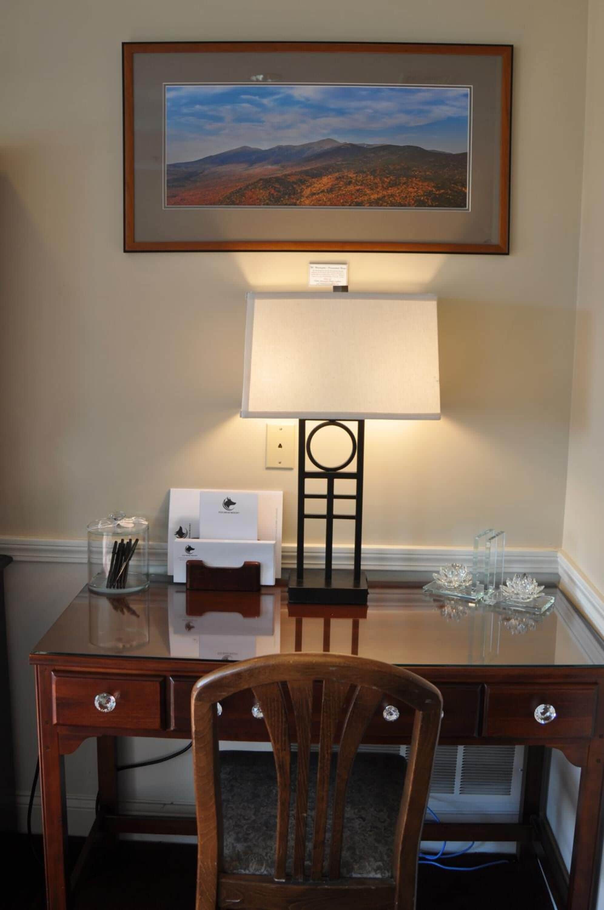 Un bureau en bois, avec un tableau représentant un paysage encadré au-dessus, une lampe et une chaise.