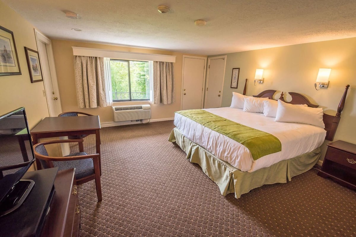 A hotel room with a bed, a desk, a chair, a window with curtains, and a wall-mounted lamp.