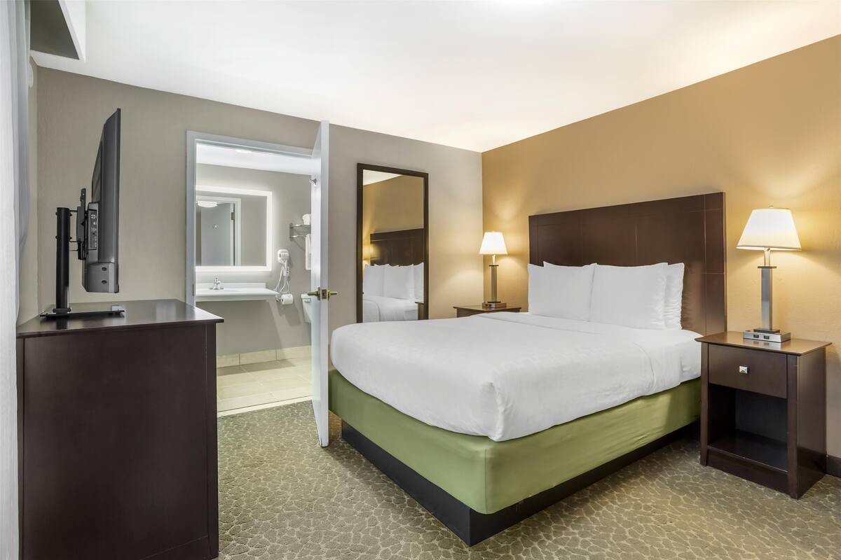A hotel room with a bed, bedside lamps, a TV, and a bathroom visible through an open door.