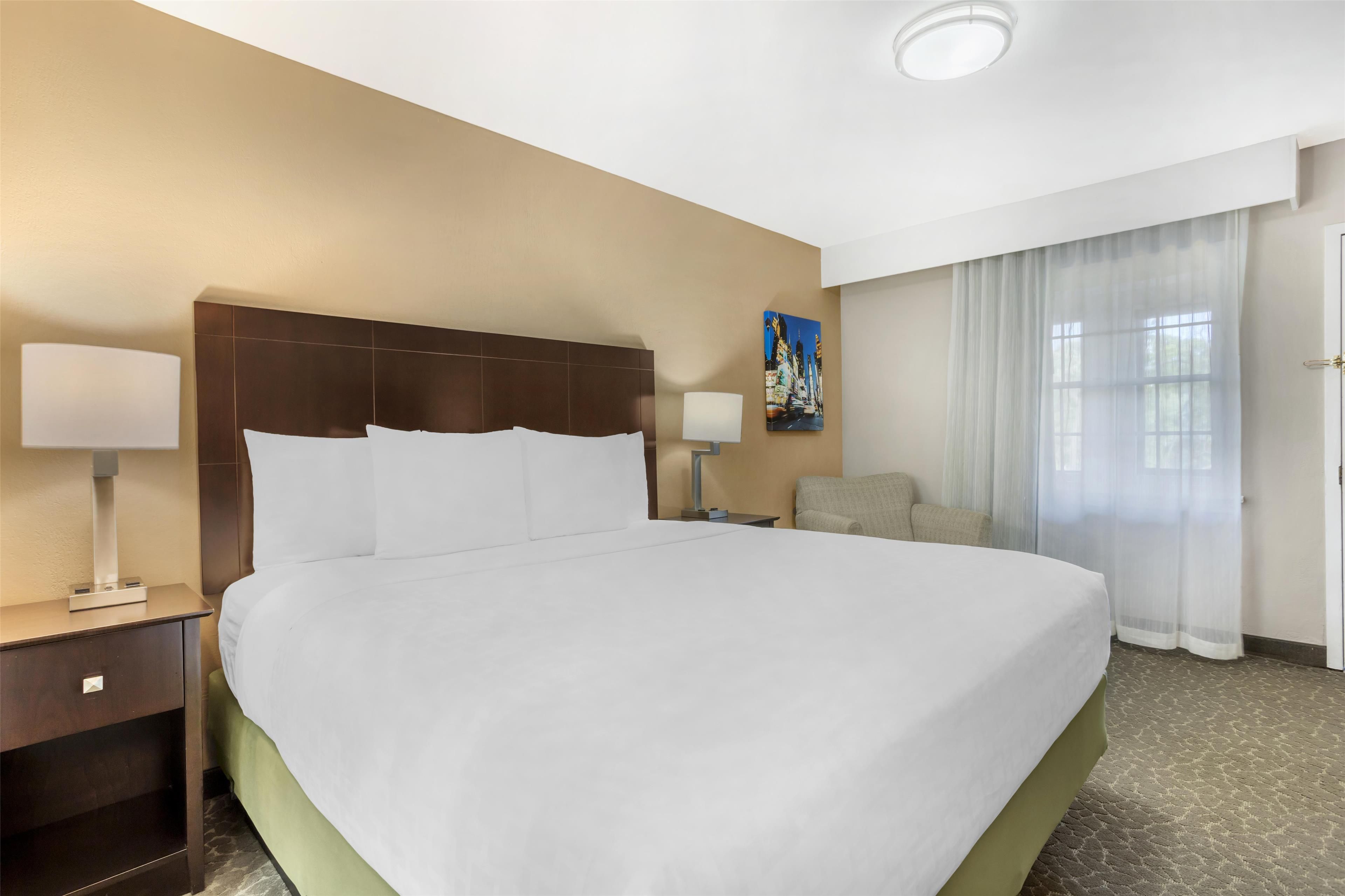 Una habitación de hotel con una cama grande, dos mesitas de noche, una silla, un cuadro en la pared y una ventana con cortinas.