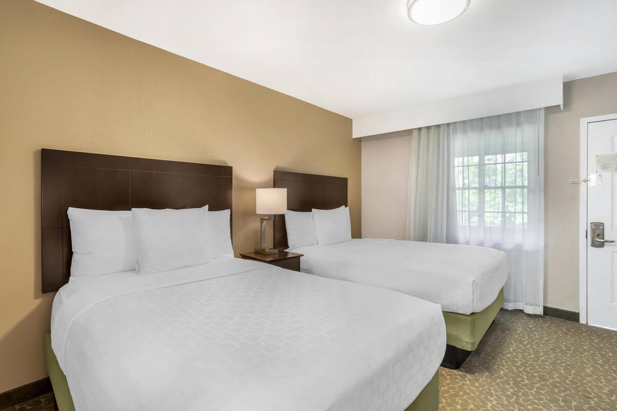 A hotel room with two beds, a nightstand with a lamp, a window with sheer curtains, and a door with a lock.