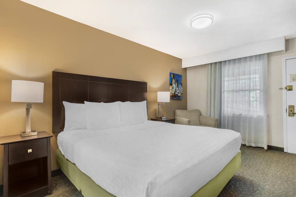A hotel room with a large bed, two bedside tables with lamps, a chair, a painting on the wall, and a window with curtains.
