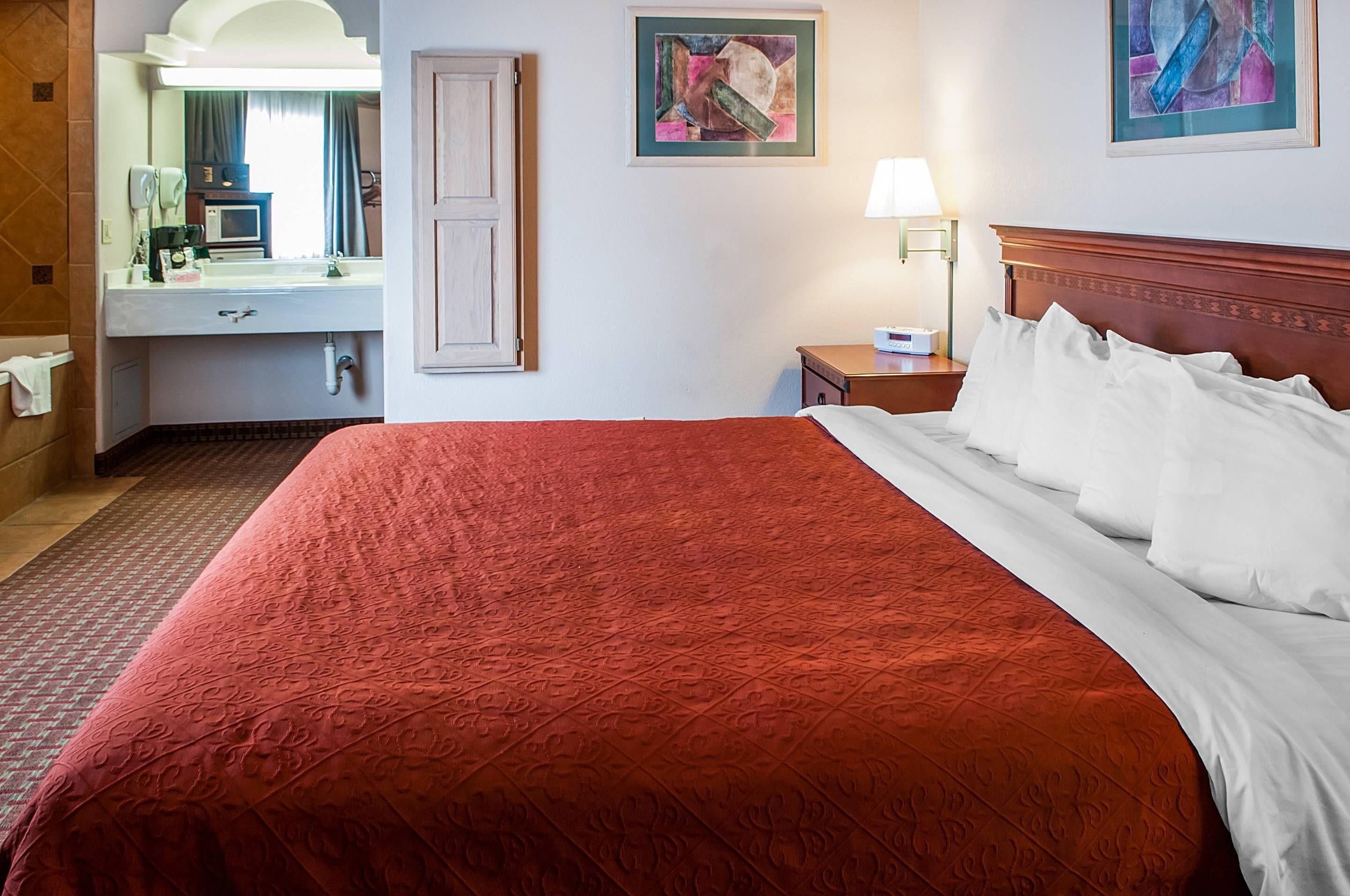 A hotel room with a large bed, a bathroom with a mirror, a sink, and a nightstand with a lamp.