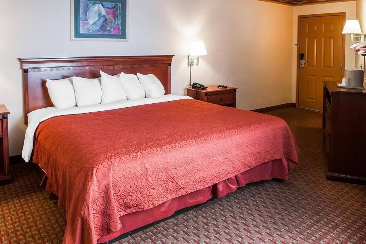 Ein Hotelzimmer mit einem großen Bett, einem Holzkopfstück, einem Nachttisch mit Lampe, einem Schreibtisch mit Telefon und einer Tür.