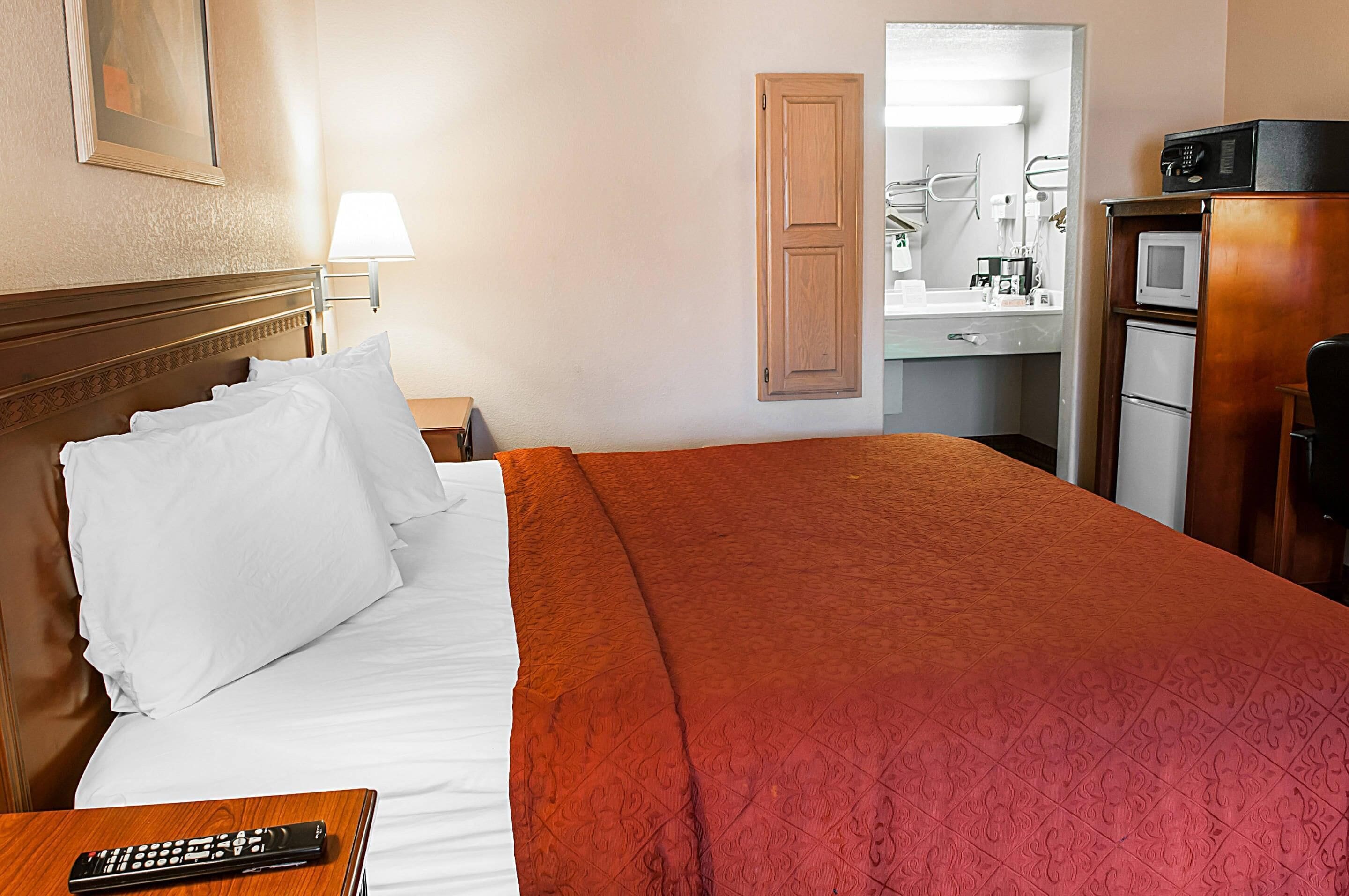 Une chambre d’hôtel comprenant un lit, une table de chevet, une télécommande, une tête de lit en bois, un miroir, un four à micro-ondes et un petit meuble de rangement.