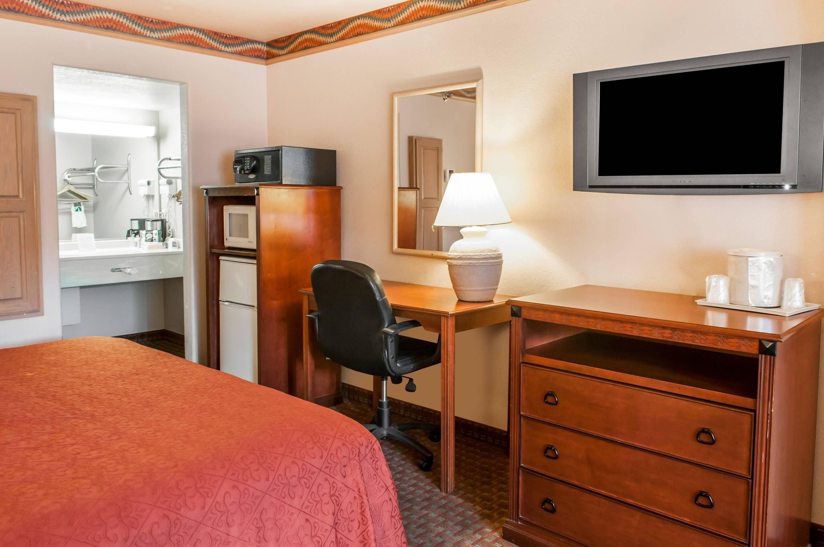 A hotel room with a bed, a desk with a chair, a television, a mirror, a lamp, a microwave, and a minibar.