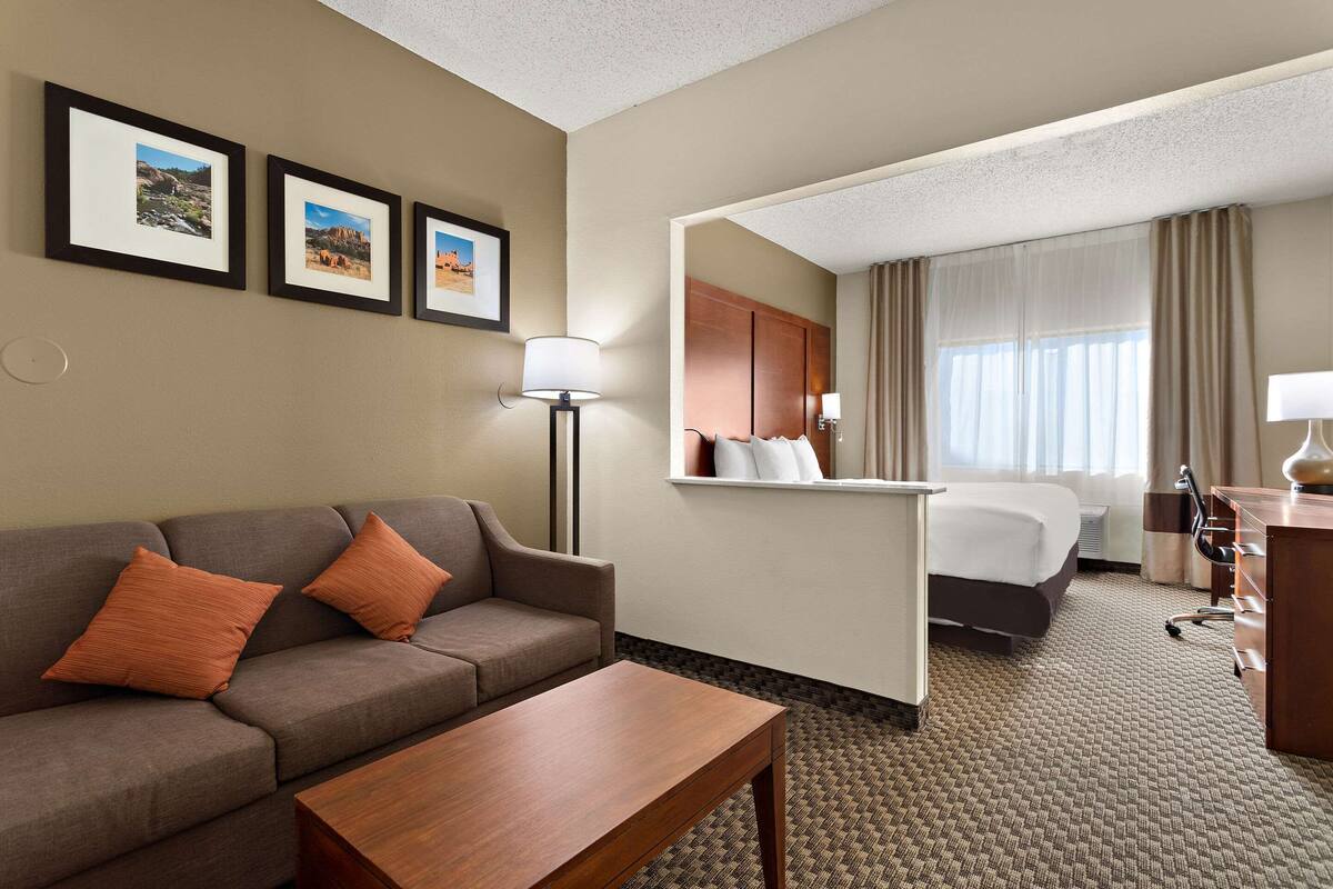 A hotel room with a sofa, a wooden coffee table, a bed, a desk, and a chair. There are framed pictures on the wall and a lamp on the desk.