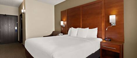 Habitación de hotel con una cama grande, cabecera de madera, mesita de noche con lámpara y piso alfombrado.