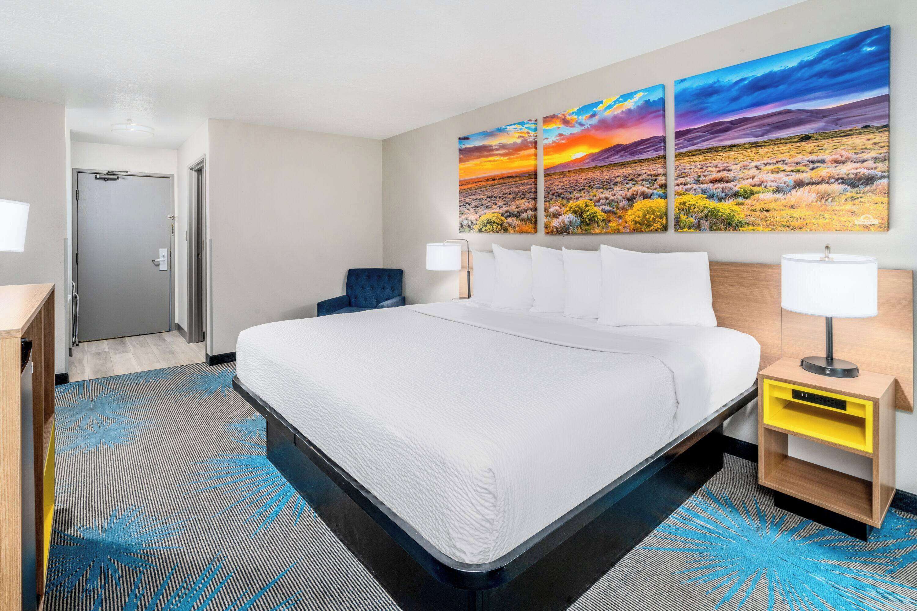 A hotel room with a large bed, a nightstand with a lamp, a wall-mounted painting, and a door leading to another room.