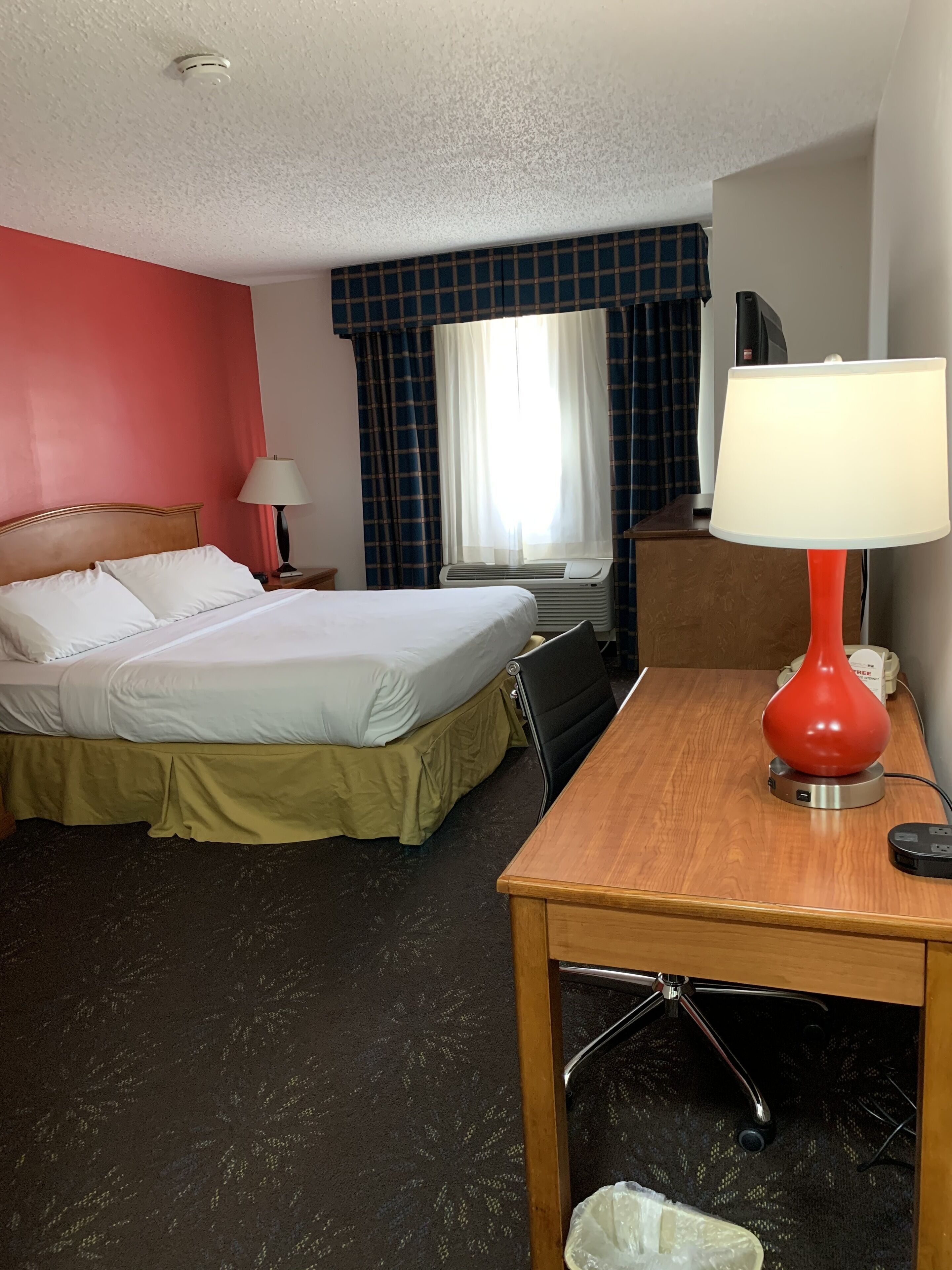 A hotel room with a bed, a desk with a lamp, a chair, a trash bin, and a window with curtains.