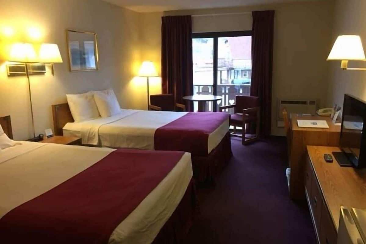 A hotel room with two beds, a desk, a chair, a TV, and a balcony with a view.