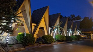 A modern building with a unique triangular architectural design, featuring multiple levels and a well-maintained garden area with trimmed bushes and a palm tree. - Tiki Resort (Lake George)