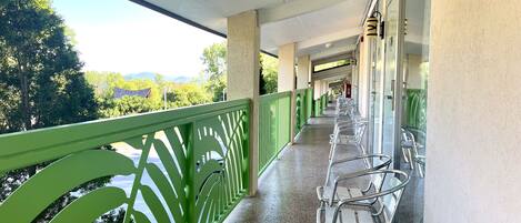 A covered outdoor balcony with metal chairs and a green railing, overlooking a scenic landscape with trees and buildings.